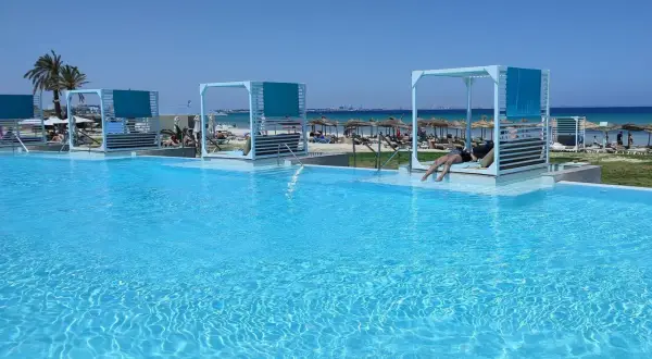 Basen z turkusową wodą, leżaki z baldachimami i plaża w tle.